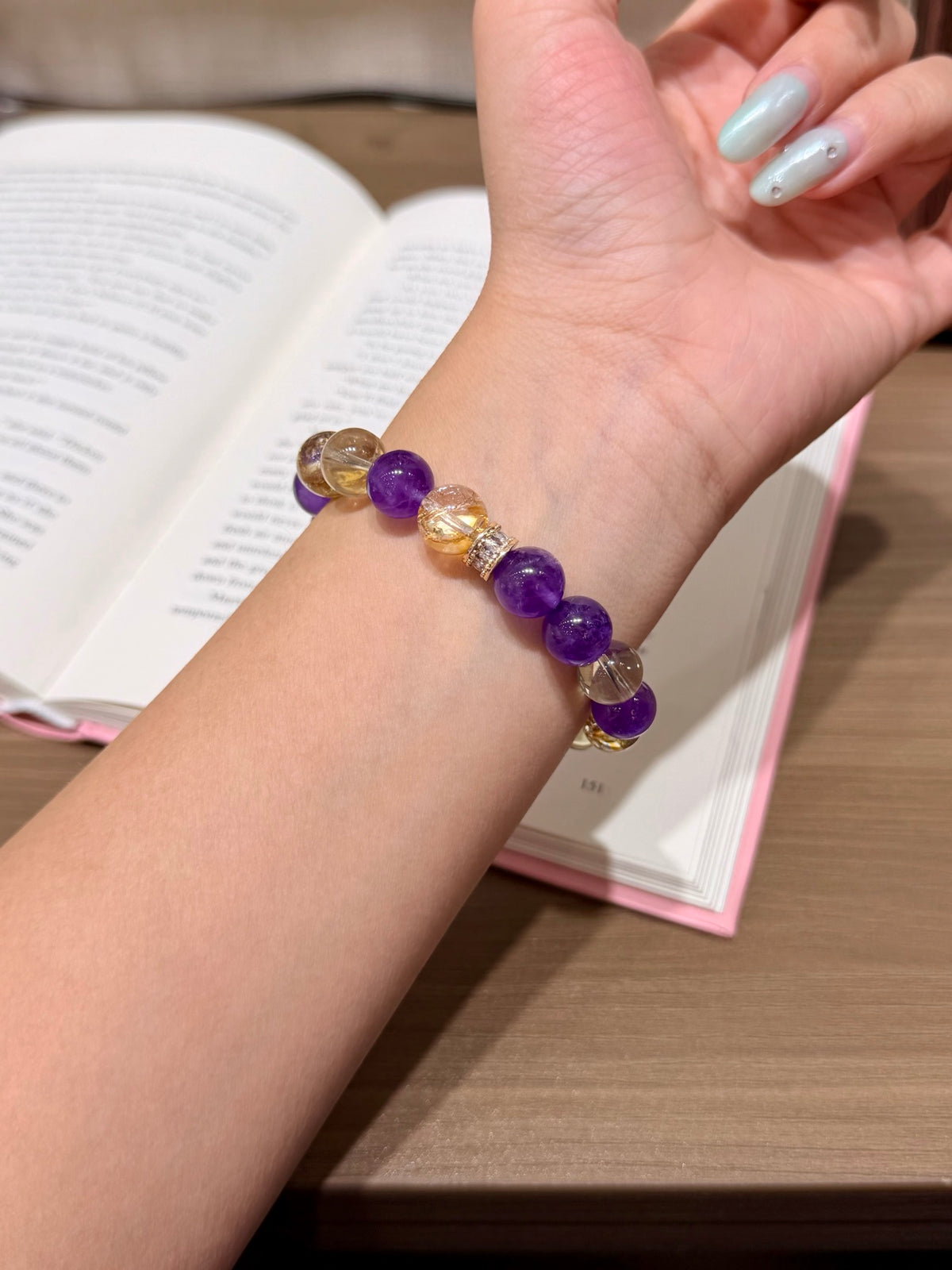 Purple and gold prologue natural crystal bracelet with amethyst and gold-foil flower crystal beads worn on a wrist with an open book in the background