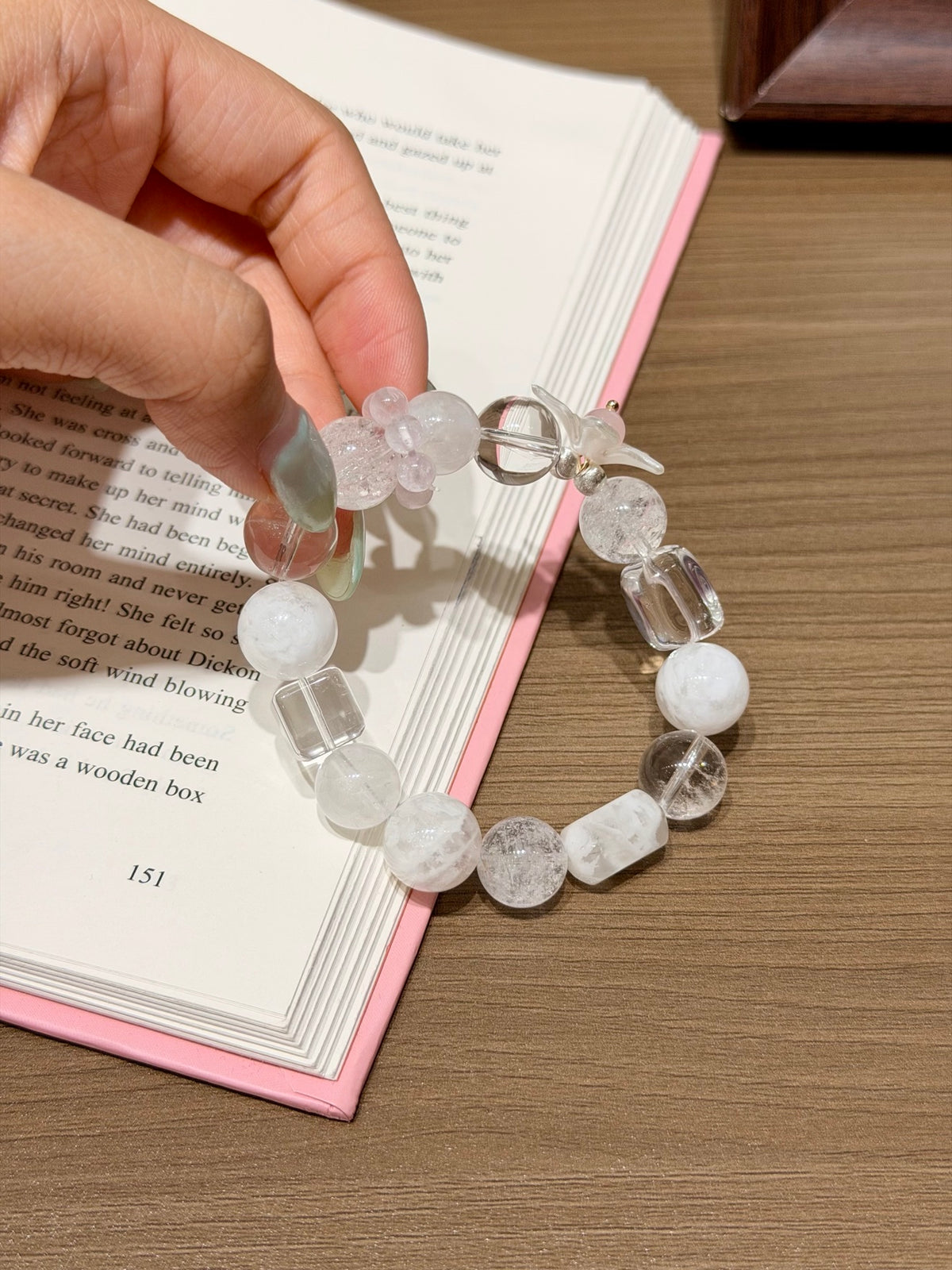 Hand holding snowfall starfield natural crystal bracelet of white phantom quartz and white agate beads with crystal cubes over an open book on a wooden table