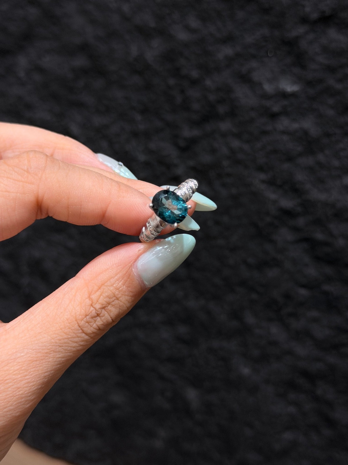 Deep teal-blue faceted topaz ring held between fingers, close-up