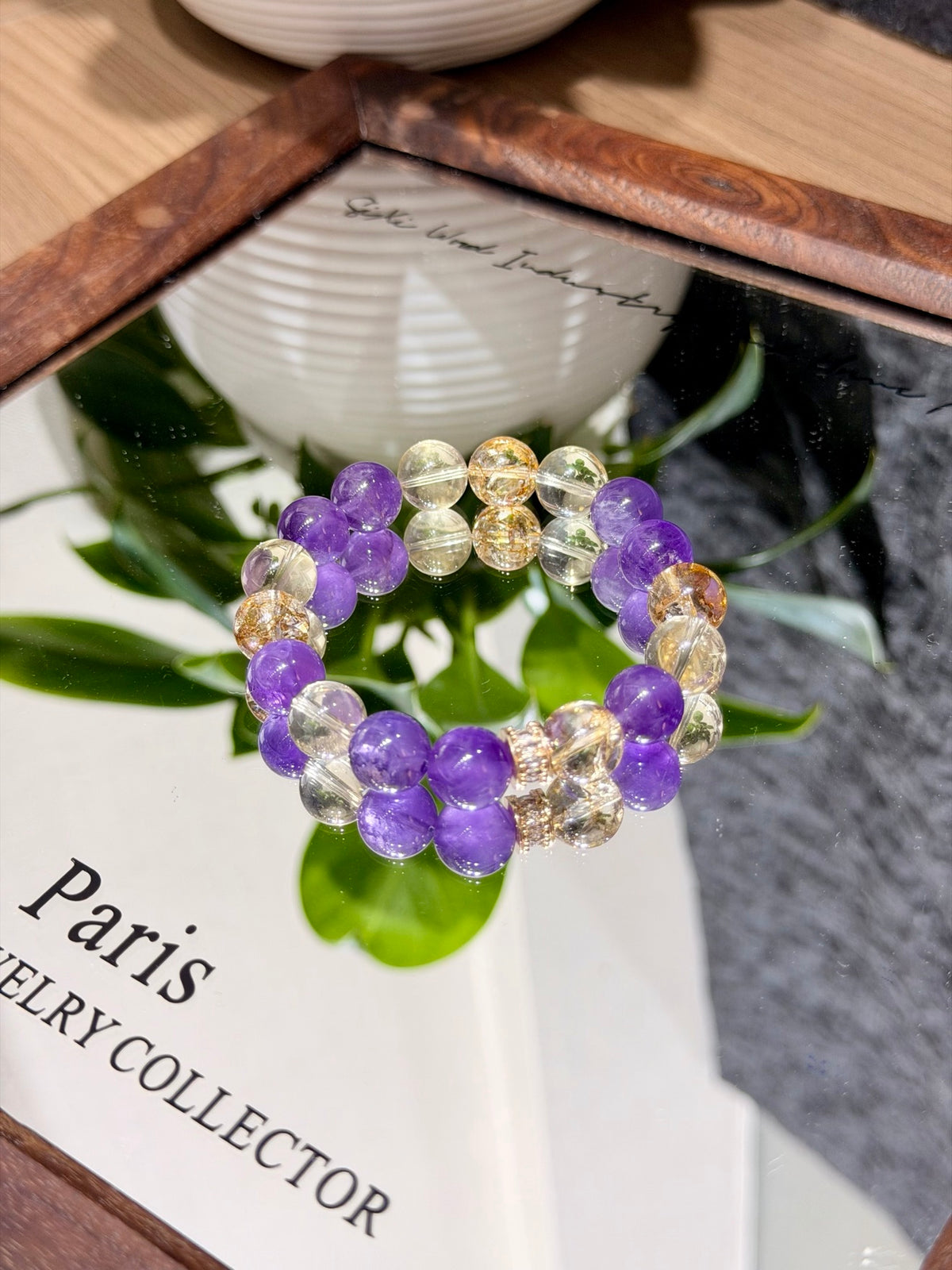 Purple and gold prologue natural crystal bracelet with amethyst and gold-foil flower crystal beads in a circle on a mirror beside a Paris Jewelry Collector book