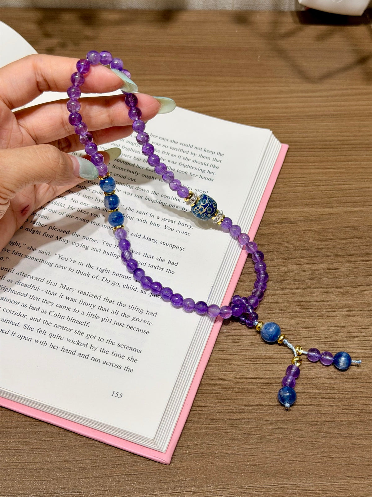 Hand holding purple amethyst double-loop bracelet with blue kyanite and patterned lacquer beads above an open book